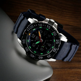 A Luminox Watches Navy SEAL Rubber, Steel, CARBONOX™ (RSC) 45 mm (SKU: XS.3253.CB) from Luminox Watches, featuring luminescent hands and markers, rests on the corner of a closed silver laptop placed on a wooden surface.