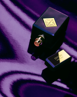 A black and gold bottle of Initio Parfums Prives Side Effect 3.0 oz EDP Perfume by Initio Perfumes, celebrated for its luxurious tobacco and vanilla notes, rests on glossy purple fabric with its cap set alongside it.