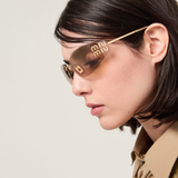 A woman with straight dark hair is seen in profile against a plain light background, wearing a beige-collared outfit and effortlessly showcasing the authentic Miu Miu Sunglasses 0MU A51S 5AK20P75 for women (UPC 8056262412367). The large brown frames feature “Miu Miu” written on the lens, highlighting the signature style of Miu Miu Sunglasses.