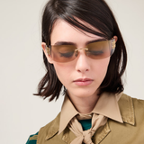A woman with short dark hair, wearing stylish Miu Miu Sunglasses 0MU A51S 5AK20P75 (UPC 8056262412367) and a beige collared shirt paired with a matching neck scarf, looks downward against a light background.
