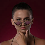A woman with slicked-back hair wears bold, angular Vysen Jaxs J-6 Unisex Sunglasses by Vysen Sunglasses and a black halter top. She gazes forward thoughtfully, her hand resting under her chin, while the deep burgundy background complements the statement shades—expertly handcrafted in Italy.