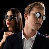 A woman and a man in dark suits wear the distinctive Vysen Nikky - NK4 Unisex Sunglasses by Vysen Sunglasses as they pose against a black background. The woman stands slightly behind with her hand on the man’s shoulder, and both display serious expressions.