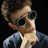 A person with curly hair, dressed in a suit and wearing distinctive Vysen Nikky - NK4 Unisex Sunglasses by Vysen Sunglasses, rests their chin on their hand, appearing thoughtful against a dark background.