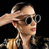 A woman with a ponytail wears Vysen Nikky - NK4 Unisex Sunglasses by Vysen Sunglasses—luxury, round, black-and-white geometric frames handcrafted in Italy—paired with a black patterned jacket. She touches the side of her face against a black background.