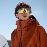 A person stands outdoors under a clear blue sky, holding a snowboard and wearing a bright orange jacket, reflective ski goggles, and sleek Oakley Meta Sunglasses 0OW8001 80010452 by Oakley Meta Sunglasses that reflect the brand’s advanced Multimodal AI integration.