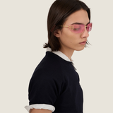 A person with straight dark hair wears Miu Miu Sunglasses 0MU A51S 5AK90Y75 and a dark short-sleeve shirt layered over a white long-sleeve top, gazing to the side against a plain light background.