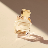 A gold-capped glass bottle of Maison Francis Kurkdjian Paris Perfumes’ Gentle Fluidity Gold 6.8 EDP Sp Unisex rests on a sunlit, textured surface, casting a delicate shadow and reflecting warm light—an elegant nod to the fragrance’s refined Oriental Vanilla notes within.
