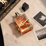 A square YSL Libre Intense 3.0 oz. EDP Women Perfume bottle with a prominent gold logo, shown with its cap removed next to two black-and-white photographs on a marble surface.