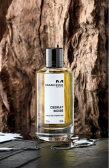 A cylindrical glass bottle of Mancera Intense Cedrat Boise 4.0 oz. EDP Women Perfume with a black cap and a white label with black text, sitting on a dark surface in front of a piece of driftwood.