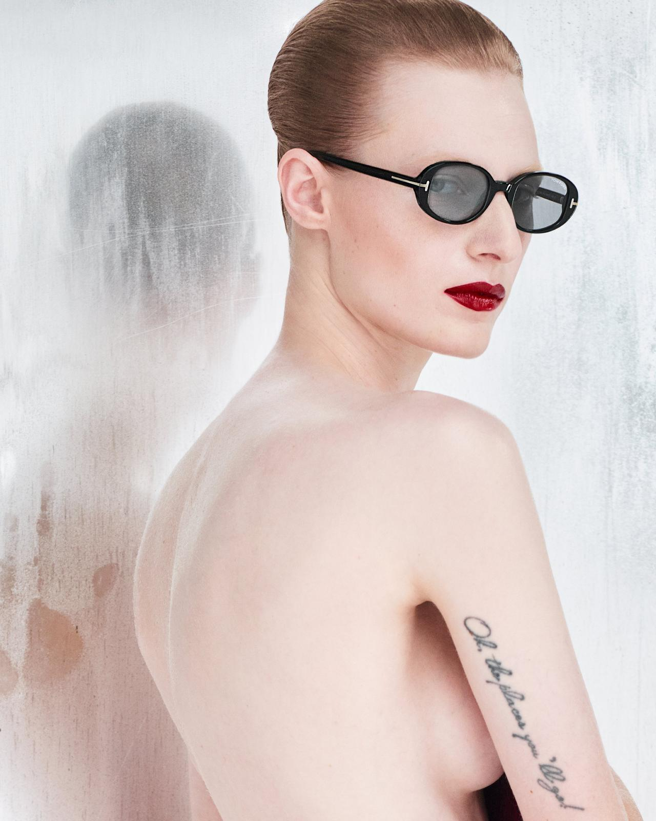 A person with slicked-back hair, bold red lipstick, and Tom Ford Sunglasses FT1364/S 52N poses with their back to the camera. A script tattoo is visible on their arm, reflected in the foggy glass behind them.