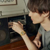 A person with straight brown hair adjusts a vintage silver boombox. Wearing a G-Shock Watches CASIO Watch A168WG-9VT and a light gray shirt, they sit at a wooden table in a softly lit room.