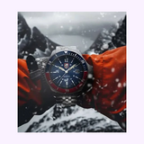 Wearing an orange jacket, a person showcases the Luminox Watches VOLITION AMERICA 'Minuteman' 42 mm XS.0914.VOL Dress Watch amid falling snow and dramatic, jagged mountain peaks in the background.