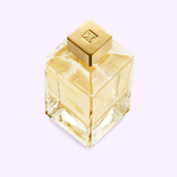 A square glass bottle filled with a light golden liquid and topped with a shiny gold cap embossed with a geometric logo is showcased against a pale background, elegantly embodying the character of Maison Francis Kurkdjian Paris Perfumes Gentle Fluidity Gold 6.8 EDP Sp Unisex—an exquisite Oriental Vanilla fragrance.