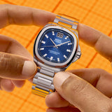 Hands holding a silver-toned Mido Watches Multifort TV Big Date Chronometer M0495671104100 with a blue checkered dial and a large date display at the twelve o’clock position.