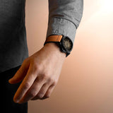 A person wearing a gray long-sleeve shirt and the Mido Watches Multifort TV Big Date M0495263608100 by Mido Watches, featuring a tan leather band, stands against a soft beige background with their hand relaxed at their side.
