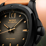 Close-up of the Mido Watches Multifort TV Big Date M0495263608100 by Mido Watches, featuring a black textured dial with gold hour markers and hands, a prominent big date display at 12 o’clock, and powered by the Caliber 80 Swiss automatic movement; partial tan leather strap is visible.