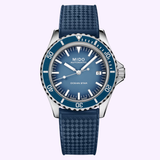 A Mido Watches M0268071104101 OCEAN STAR TRIBUTE Swiss automatic watch featuring a blue dial, stainless steel bezel, silver-tone hour markers, a date display at 3 o’clock, and a textured rubber strap is displayed against a light purple background.
