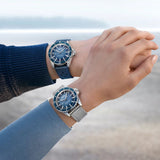 Two people wearing matching silver and blue wristwatches, specifically the Mido Watches M0268071104101 Ocean Star Tribute, hold hands outdoors with a softly blurred natural background.