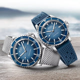 Two blue Mido M0268071104101 Ocean Star Tribute watches by Mido Watches, each with a stainless steel case, are displayed on a stone surface by the sea—one adorned with a mesh band and the other with a textured blue strap. Both timepieces feature striking dials and silver hour markers, embodying the enduring precision of Swiss automatic craftsmanship.