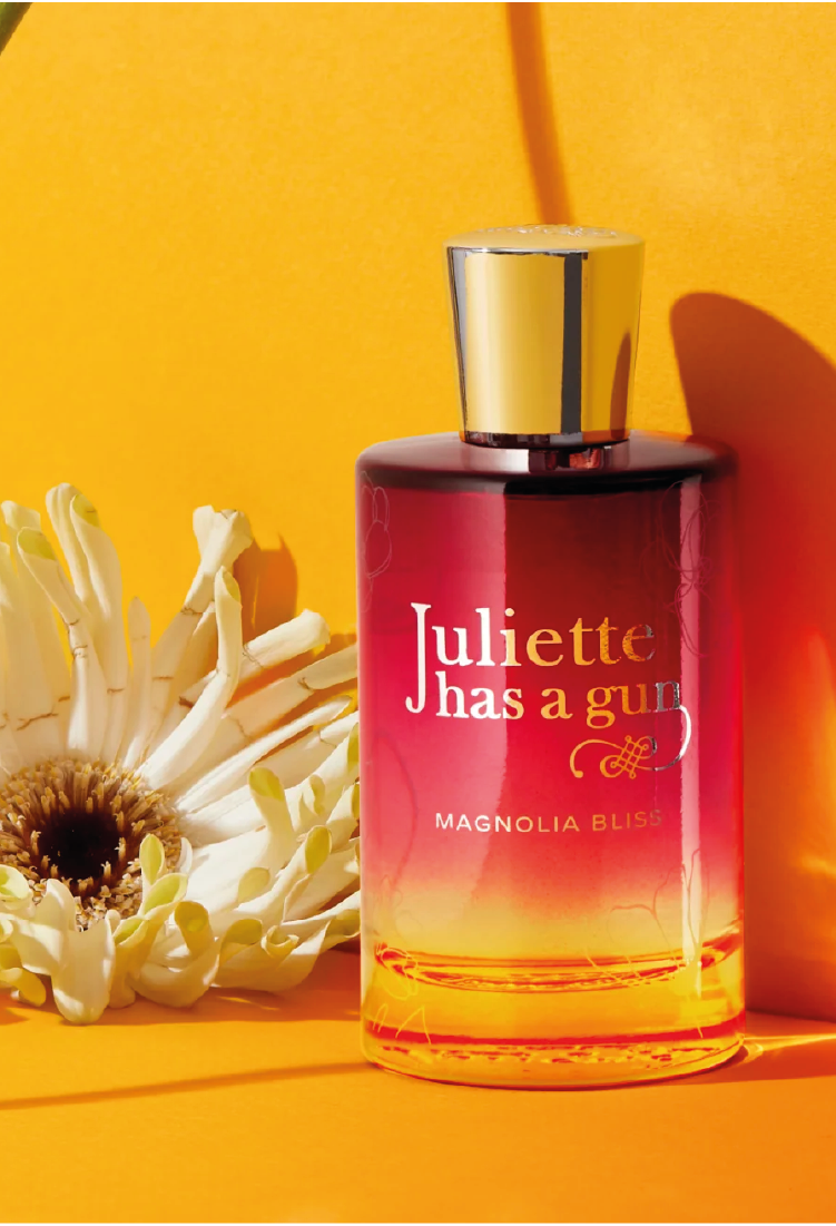 A bottle of Juliette Has a Gun Magnolia Bliss perfume with a gold cap is placed on a bright yellow surface beside a white flower, with yellow light casting soft shadows.