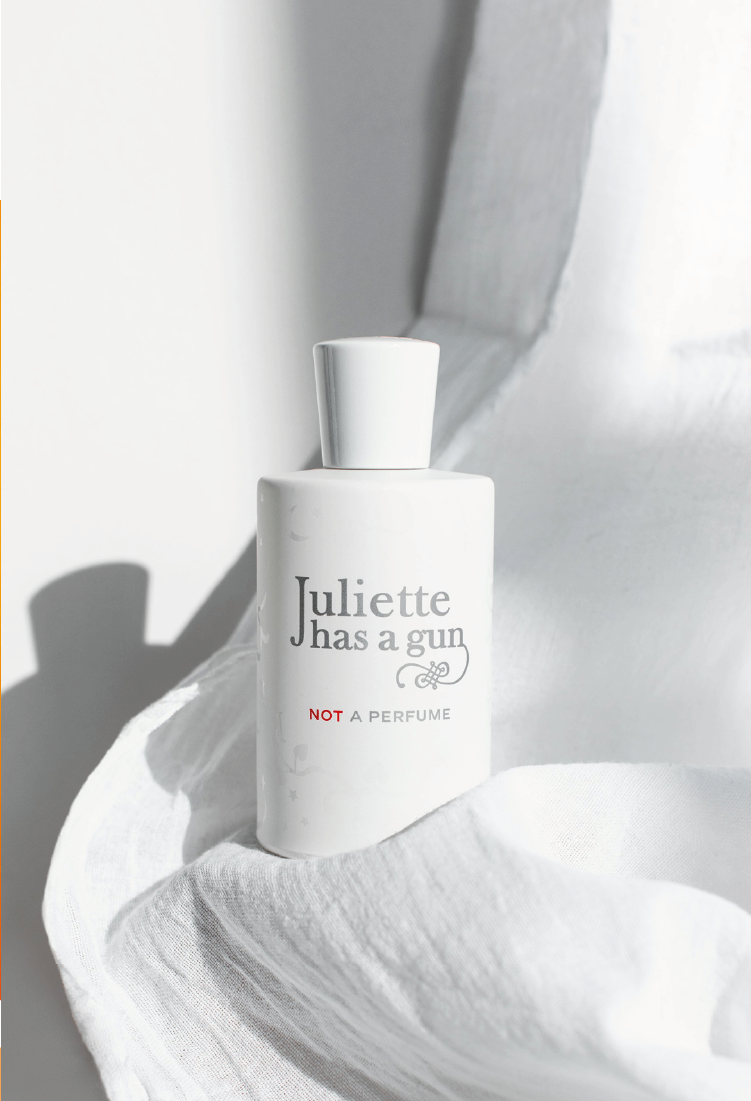 A white bottle labeled Juliette has a gun NOT A PERFUME stands on soft, white fabric, with sunlight casting gentle shadows in the background.