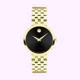 A gold-tone women’s watch featuring a black face with a minimalist design and a signature single dot at the 12 o’clock position. The Movado Watches brand name appears near the 6 o’clock mark, highlighting the elegant craftsmanship of the Museum Classic model (0607847) from this iconic Swiss brand.