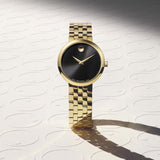 The Movado Watches 0607847 Museum Classic, a gold-tone women’s watch from Movado Watches, showcases a sleek black minimalist dial highlighted by the iconic single gold dot at 12 o’clock. Displayed upright against a patterned backdrop and casting an elegant shadow, this Swiss timepiece is finished with a sophisticated gold link bracelet.