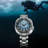The Seiko Luxe Watches PROSPEX BUILT FOR THE ICE DIVER U.S. SPECIAL EDITION SPB263 is showcased with its striking blue textured dial, stainless steel bracelet, and rotating bezel, while a scuba diver swims underwater in the background.