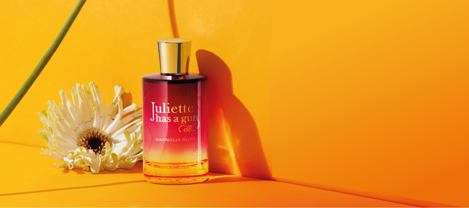 A Juliette Has a Gun Magnolia Bliss perfume bottle sits on a vibrant orange surface beside a white daisy, with soft shadows and a bright, warm background.