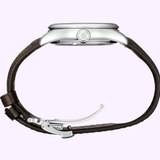 A side view of the Seiko Luxe Watches Presage Green Enamel Limited Edition SPB111 men’s watch, featuring a round silver case, an S-engraved crown, and a brown leather strap with a metal deployment clasp, set against a light purple background.