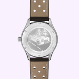 The Frederique Constant Vintage Rally Healey Automatic FC-301HGRS5B26 features a brown perforated leather strap, racing green dial, and a stainless steel back engraved with a classic car on a pine-lined road.
