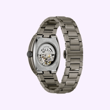 The Bulova Maquina 98A321 by Bulova Watches is a stainless steel automatic men’s watch with an exhibition case back and silver metal link bracelet, elegantly showcased against a light purple background.