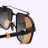 Close-up of Balmain Sunglasses BRIGADE-V BPS-142E-56 Unisex showing perforated gold temple accents and dark lenses against a light background. A modern, standout accessory from Balmain Sunglasses.