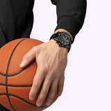 A person in a black long-sleeve shirt wears the Tissot Watches Supersport NBA Special Edition T1256173706700 and holds an orange basketball against a light background.