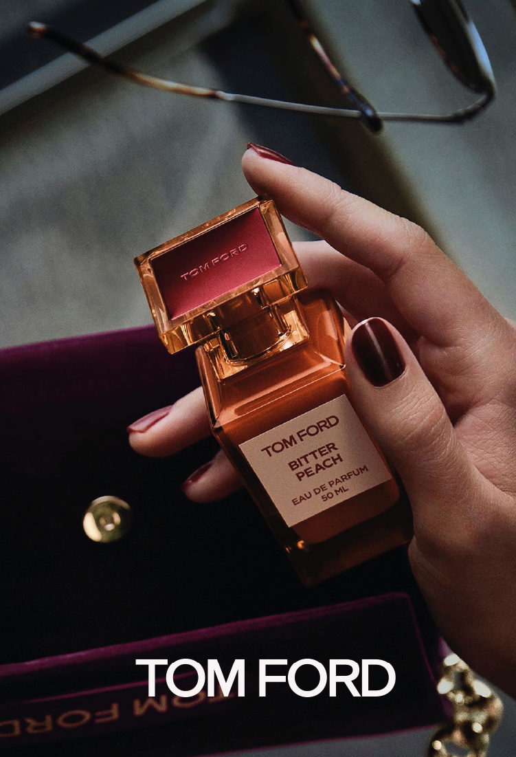 A hand with dark red nail polish holds a Tom Ford Bitter Peach perfume bottle above an open velvet box, with sunglasses in the background. The Tom Ford logo is visible at the bottom of the image.