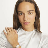 A woman with long brown hair, wearing a white satin top and the Tory Burch Watches THE OVAL TBW6037 on her wrist, looks at the camera with a neutral expression, her hand resting on her shoulder.