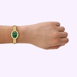 A hand wearing the Tory Burch Watches THE OVAL Women TBW6038, featuring a gold-tone stainless steel linked band and a striking green malachite dial, is shown against a light purple background.