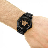 A person wearing a Versace VERD00518 Palazzo Empire 43 mm men's watch featuring a black metal band, gold Medusa head on the black dial, gold hour markers, and precise Swiss Quartz movement from Versace Watches.