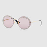 Gucci GG0899S 001 61 Women Sunglasses feature round frames with pale pink lenses, gold metal arms tipped in black, and sparkling crystal embellishments. Inspired by chic Gucci style. Displayed on a plain light gray background.