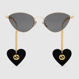 The Gucci GG0977S 001 57 Women Sunglasses feature a gold cat-eye frame, dark lenses, and temples with interchangeable gold chains ending in black heart-shaped charms with the iconic gold Gucci logo—perfect for fans of chic Gucci sunglasses.