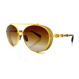 The Balmain BPX-111A-60 Brigade-II Unisex Sunglasses feature gold rims, brown gradient lenses, side shields, and black and gold arm detailing. These designer Balmain Sunglasses are shown against a white background.