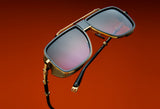 The Balmain O.R. BPS-104C-59 Unisex Sunglasses by Balmain Sunglasses feature gold frames, purple-tinted lenses, distinctive gold accents, and black arms, shown against a bold red-orange background for a modern look.
