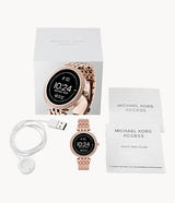 The Michael Kors MKT5128 Darci Gen 5E Rose Gold Stainless Steel Women’s Smartwatch is shown with its box, USB charger, and two manuals on a white background. The digital clock display appears on the watch screen.