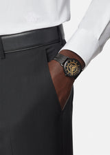 A person in black dress pants and a white shirt with an embroidered logo has their hand in their pocket, showcasing the Versace VEJ400621 V Essential 40 mm Men Watch by Versace Watches with a stainless steel mesh band.