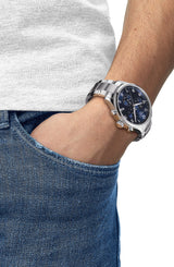 A person wearing a swatch group Tissot T1166171103700 Chrono XL Classic Men’s Watch with a stainless steel strap and black dial, paired with a white t-shirt, has their hand casually in the front pocket of blue jeans.