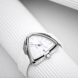 A close-up of the Hamilton H24505311 Ventura Elvis80 Auto unisex watch by Hamilton Watches, featuring a unique triangular face, silver bezel, blue hands, date at 3 o’clock, H-10-S automatic movement, and a white silicone strap.
