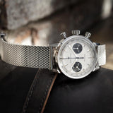 The Hamilton H38429110 American Classic Intra-Matic Chronograph by Hamilton Watches features a silver case, white dial with black subdials, and mesh metal strap, shown on dark leather near a brick wall.