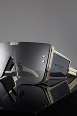 Close-up of Balmain BPS-102F-146 Wonder Boy LTD Unisex Sunglasses by Balmain Sunglasses, featuring large angular dark lenses and metallic accents, displayed on a glossy black surface with a soft gray background.