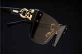 Close-up of Balmain BPS-106B-150 Armour Unisex Sunglasses in black with gold-accented frames and hinges. The tinted brown lenses and interlocking circular shapes showcase these iconic Balmain Sunglasses against a dark background.