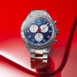 The Tissot T1316171104200 PRS 516 CHRONOGRAPH Men’s Watch by Tissot Watches features a stainless steel case, blue dial with three subdials, and tachymeter bezel, shown against a glossy red background.